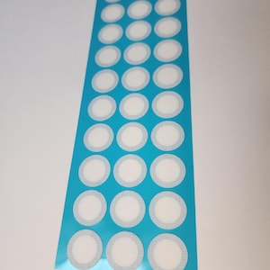 Peut inclure: Une feuille de pansements adhésifs ronds et blancs avec un support bleu clair. Les pansements sont disposés en rangées et en colonnes. Le support est de couleur bleu vif.