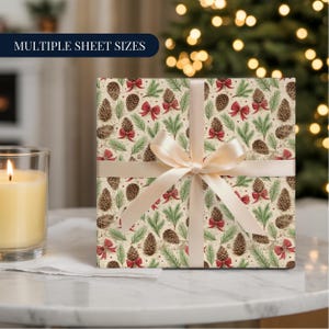 Puede incluir: Un regalo envuelto con una cinta color crema. El papel de regalo presenta un patrón de piñas, lazos rojos y agujas de pino verdes sobre un fondo crema. El texto "MULTIPLE SHEET SIZES" está en un rectángulo azul marino. Una vela encendida está a la izquierda.