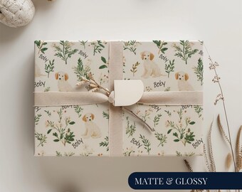 Può includere: Un regalo avvolto con carta color crema con un motivo di cuccioli di golden retriever ed elementi botanici. È legato con un nastro beige e un'etichetta. Le parole "Matte & Glossy" sono in basso.
