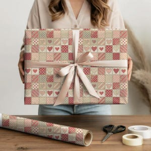 Puede incluir: Un regalo envuelto en papel con un diseño de patchwork de corazones y lunares en tonos rojos, rosas y beige. Está atado con una cinta rosa claro. También se ve un rollo de papel de regalo a juego.