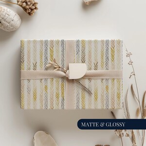 Puede incluir: Una caja de regalo envuelta con papel con un patrón de flechas verticales verdes, grises y beige. Está atada con una cinta y etiqueta crema. Las palabras "MATTE & GLOSSY" están en un rectángulo azul marino.