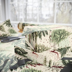 Puede incluir: Cajas de regalo envueltas con papel festivo con un diseño de bosque en colores verde, marrón y crema. La escena incluye tijeras, cinta y papel adicional, lo que sugiere una festividad u ocasión especial. El papel tiene un patrón repetido.