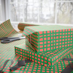 Puede incluir: Dos cajas de regalo envueltas en papel a cuadros verde y rojo están apiladas. Un rollo de papel de regalo a juego y unas tijeras son visibles en el fondo. Una cinta negra está colocada sobre el papel de regalo. Envolviendo regalos.