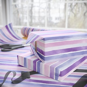 Puede incluir: Dos cajas de regalo envueltas en papel a rayas morado, lavanda y blanco. Se ven un rollo de papel de regalo a juego y unas tijeras. Una cinta negra está drapeada en la parte inferior, lo que sugiere un regalo terminado.
