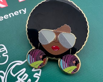 May include: A gold-toned enamel pin featuring a cartoon woman with an afro hairstyle, wearing sunglasses and large colorful earrings. The woman has brown skin and red lips.