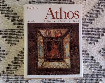 May include: A book titled "Athos" by Paul Huber, featuring an image of religious art. The book's cover has text in English and German. The book is resting on a white and gray patterned rug.