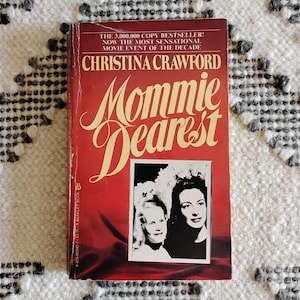 May include: A paperback book titled "Mommie Dearest" by Christina Crawford. The cover is red with the title in large, cursive, gold lettering. A black and white photo of two women is in the center. The book is on a white and gray patterned surface.