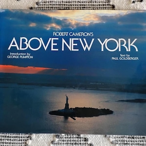 May include: A book cover with a photograph of New York City at dusk. The title "ABOVE NEW YORK" is in large white letters. The Statue of Liberty is visible in the water. The book is by Robert Cameron.
