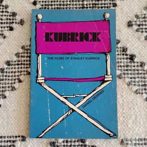 May include: A book cover with a director's chair design. The word "KUBRICK" is in black block letters on a pink background. Below, the text reads "THE FILMS OF STANLEY KUBRICK" and "DANIEL DEVRIES" on a blue background.