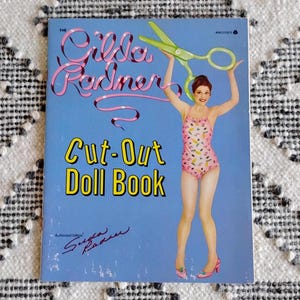 May include: A vintage "Gilda Radner Cut-Out Doll Book" cover. The cover features a woman in a pink swimsuit holding green scissors. The title is in yellow and the author's signature is at the bottom. The background is light blue.
