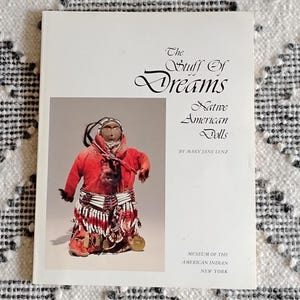 May include: A book cover featuring a Native American doll in a red outfit. The title "The Stuff of Dreams" is displayed in elegant script. The book is by Mary Jane Lenz and published by the Museum of the American Indian, New York.