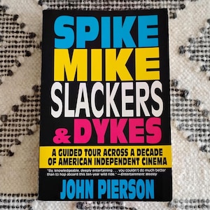 May include: A book titled "SPIKE MIKE SLACKERS & DYKES" with a black cover and colorful text. The title is in blue, yellow, white, and pink. The book is about American independent cinema and is written by John Pierson.