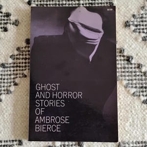 May include: A paperback book titled "Ghost and Horror Stories of Ambrose Bierce" with a dark cover featuring a shadowy figure. The book has a price tag of $2.00 in the upper right corner. The book is set against a patterned white and gray background.