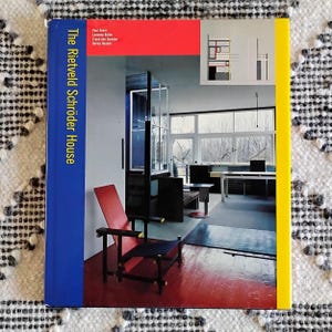 May include: A book titled "The Rietveld Schröder House" with a blue spine and a red, blue, and yellow cover. The cover features an interior photo with a red chair and a black and white patterned rug.