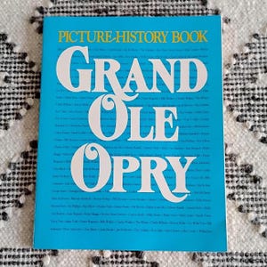 May include: A picture history book titled "Grand Ole Opry" with a bright blue cover. The title is in large white letters, with smaller text of names and phrases. The book is a vintage item.