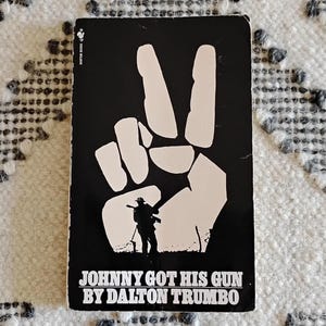 May include: A black and white book cover for "Johnny Got His Gun" by Dalton Trumbo. The cover features a white hand making a peace sign, with a silhouette of a soldier. The title and author's name are in white.
