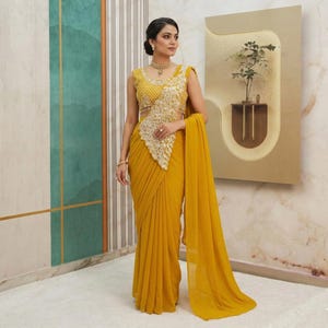 Pode incluir: Um sari amarelo mostarda com uma blusa sem mangas a condizer. A blusa apresenta bordados dourados intrincados no corpete. O sari drapeia-se elegantemente, com um pallu fluido. O traje é usado com joias douradas.