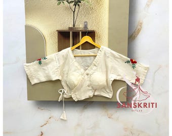 May include: Cream-coloured wrap blouse with floral embroidery on the sleeves. The blouse has a V-neck, button closure, and tie closure at the waist. The blouse is hanging on a yellow hanger.
