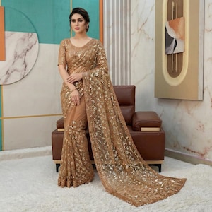May include: A gold-colored saree with intricate sequin detailing. The saree features a flowing design with a matching blouse. The fabric appears to be a sheer material, creating a shimmering effect. The model is standing in front of a brown leather chair.