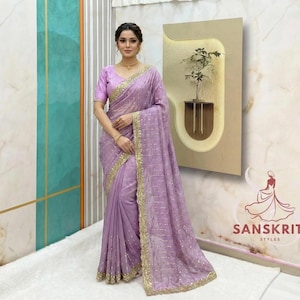 Puede incluir: Un sari lavanda con adornos y adornos dorados. El sari se usa con una blusa a juego. El fondo incluye un panel de pared decorativo y un logotipo que dice "Sanskriti Styles."