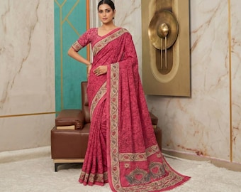 May include: A woman wearing a vibrant pink saree with a floral pattern. The saree features a detailed border with intricate designs in beige and brown. The woman is standing in front of a decorative wall with a brown leather chair.