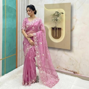 May include: A light pink saree with intricate white floral embroidery along the borders and edges. The saree features a sheer, flowing fabric and is worn by a person. The outfit includes a matching blouse and is accessorized with gold jewelry.