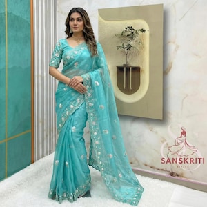 May include: A light blue sari with intricate silver embroidery and a sheer overlay. The sari features a detailed floral pattern along the borders and sleeves. The image shows a woman wearing the sari, showcasing its elegant design.