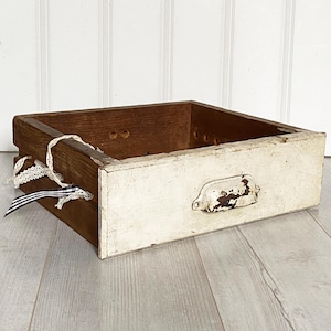 May include: A vintage wooden drawer with a distressed white front and a brown wooden frame. The drawer has a metal handle and is decorated with lace and a striped ribbon. The drawer is on a light grey wooden surface.