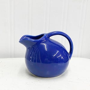 May include: A cobalt blue ceramic pitcher with a rounded body and a curved handle. The pitcher is sitting on a white surface.