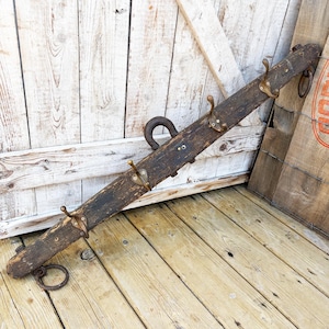 Peut inclure: Une longue poutre en bois brun foncé patiné reconvertie en porte-manteau mural. Elle comporte quatre crochets en métal doré fixés sur le devant, ainsi que deux grands anneaux en fer rouillé aux extrémités et une boucle centrale en fer.