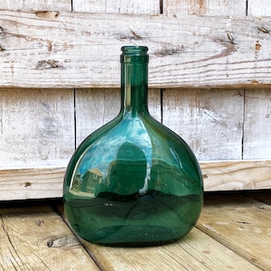 May include: A large, green glass bottle with a rounded body and a long neck. The bottle is sitting on a wooden surface. The bottle's surface reflects the surrounding environment. The bottle is empty.