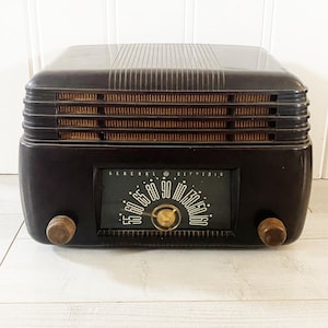 May include: A vintage General Electric radio in a dark brown, rectangular design. The radio features a speaker grill, a tuning dial with the text "GENERAL ELECTRIC", and two wooden knobs. A classic piece of home decor.