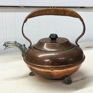 May include: A small copper teapot with a woven handle, a silver spout, and three silver feet.