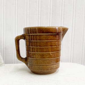 May include: A brown glazed ceramic jug with a wide mouth and a handle. The jug has a rustic, vintage look and is decorated with horizontal lines.