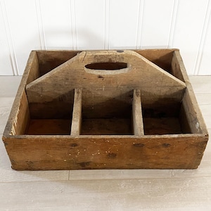 May include: A rustic, wooden carrier with a handle and four compartments. The carrier is made of weathered wood with visible grain and knots, and shows signs of age and use. The handle has a large oval opening for easy carrying.