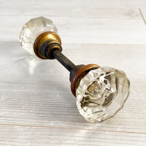 Peut inclure: Un bouton de porte vintage avec des boutons en verre clair et facetté et des garnitures en laiton. Le bouton de porte est posé sur un fond en bois clair, mettant en valeur son design antique. Le design du bouton est un style classique et intemporel.