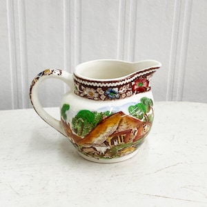 Peut inclure: Un petit pot à crème en céramique blanche avec un motif floral brun, vert et rouge. Le pot à crème représente une scène de cottage avec un toit de chaume et un champ vert.