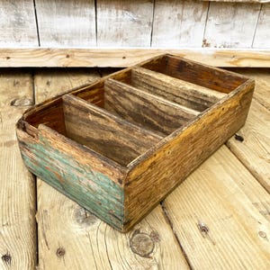 May include: A vintage wooden box with four internal compartments. The box is constructed from aged wood, displaying natural grain patterns and knots. One end of the box has faded teal paint, contrasting with the aged wood. The box is placed on a wooden surface.