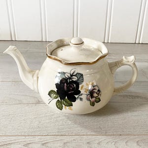 May include: Cream ceramic teapot featuring a black rose floral design. The teapot has a gold trim around the lid, a curved spout, and a handle. The floral design includes black and pink roses with green leaves.