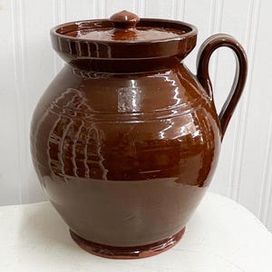 May include: A large, brown ceramic crock with a lid and a handle. The crock has a glazed finish and is decorated with a simple design.