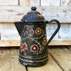 May include: Black vintage coffee pot with a lid and handle. The pot is decorated with red and gold floral and bird designs. The pot is sitting on a wooden surface.