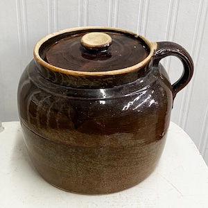May include: A brown glazed ceramic crock with a lid and a single handle. The crock has a rustic, vintage look.