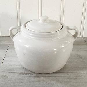 May include: A white ceramic lidded pot with two loop handles. The pot has a rounded body and a lid with a small knob. The background is a white vertical panel and a gray wooden surface.