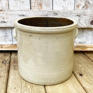 Peut inclure: Un grand pot en grès blanc cassé avec un intérieur brun foncé. Le récipient cylindrique a deux petites poignées et un bord légèrement évasé. Le texte "EL FABBRIC" est estampé sur le côté. Le pot est posé sur une surface en bois.