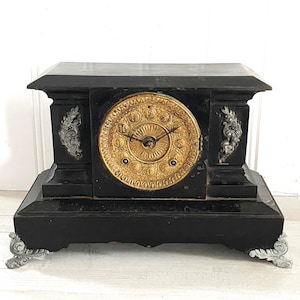 May include: Black antique mantel clock with a gold clock face and silver accents. The clock has a rectangular shape with ornate details and sits on four decorative feet. The clock measures approximately 30 cm in height.