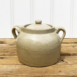 May include: A speckled, beige ceramic pot with a lid and two curved handles. The pot is round with a slightly textured surface. The lid has a small knob. The pot is sitting on a wooden surface.