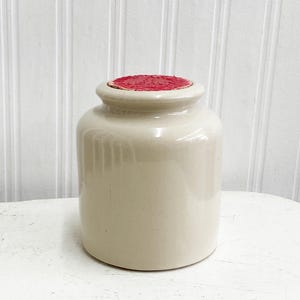 Peut inclure: Un pot en céramique blanche avec un couvercle rouge. Le pot a un design côtelé et est parfait pour ranger de petits objets.