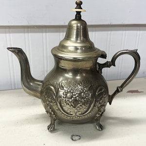 Puede incluir: Una tetera de plata con un diseño floral adornado y tres patas. La tetera tiene una tapa con un pequeño pomo en la parte superior.