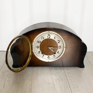 May include: A vintage wooden mantel clock with a glass cover. The clock face is white with black Roman numeral hour markers and black hands. The clock is in a dark wood case with a curved top.
