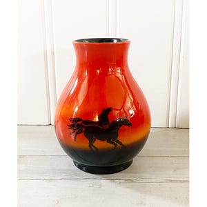 May include: A glossy, orange ceramic vase with a black base and rim. The vase features a silhouette of a horse with wings, set against a gradient of orange and black. The vase is a decorative item.
